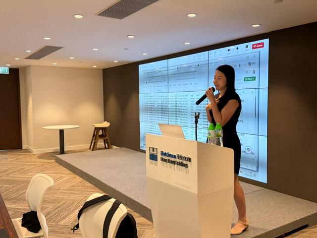和記電訊講台上，Eva 手持咪高峰，身後大螢幕顯示工作檔案